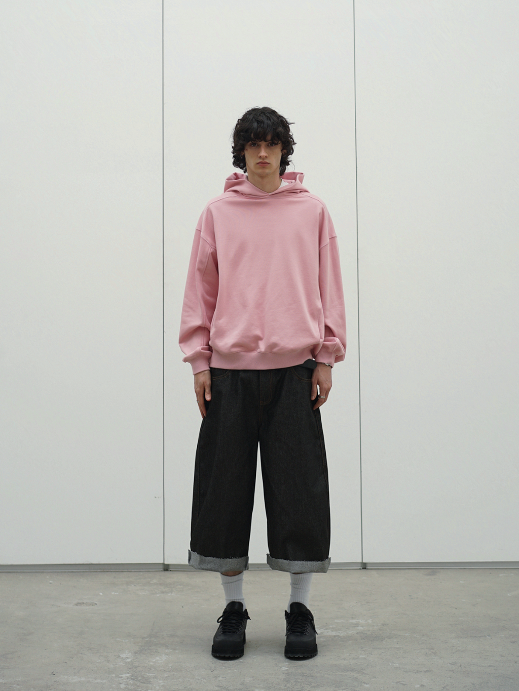 Person wearing a pink hoodie and black cropped jeans standing against a white wall.