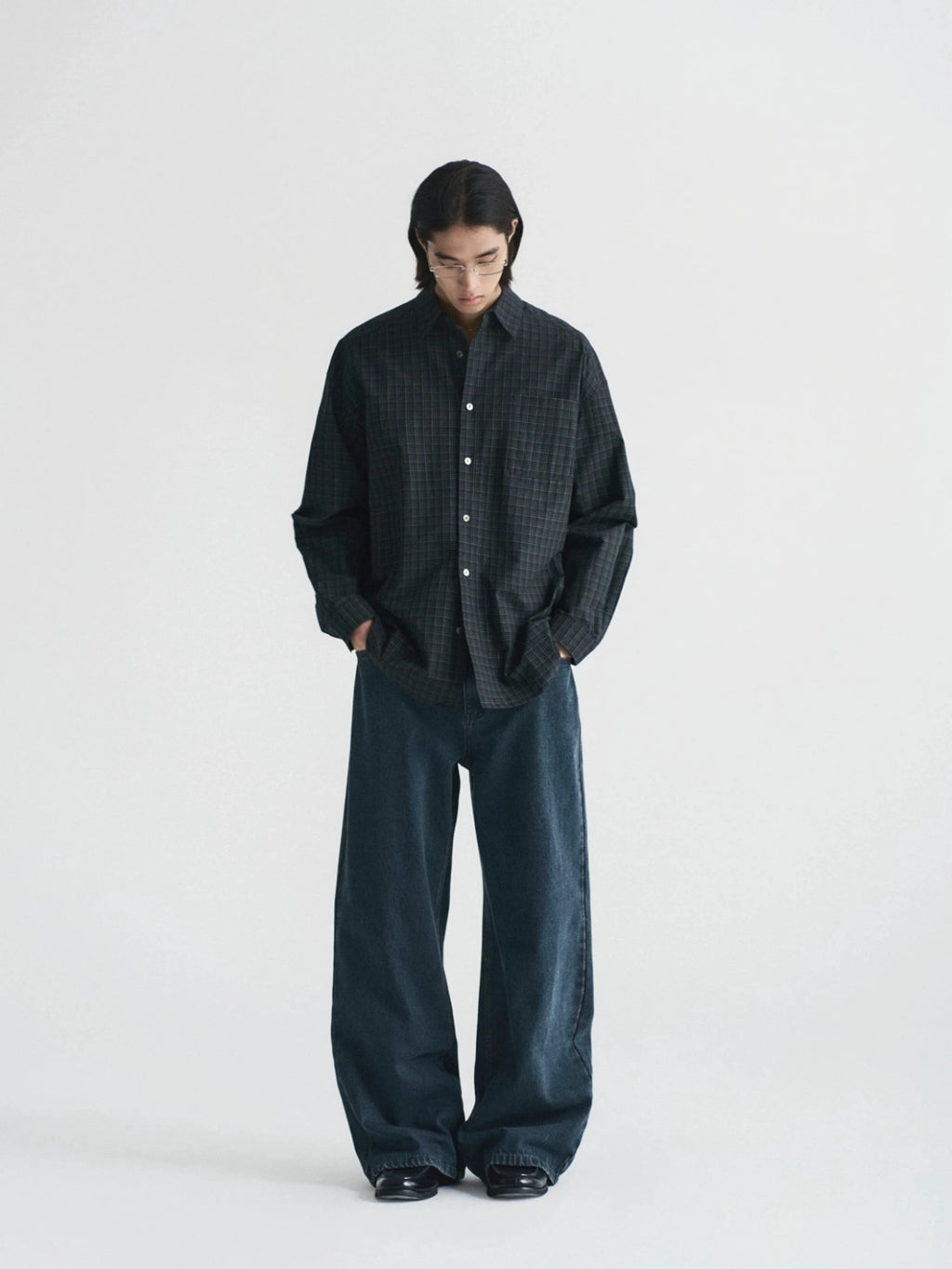 A model stands against a plain light gray studio background wearing a dark oversized button-front shirt with a subtle grid pattern and very wide-leg dark trousers from Inknots.