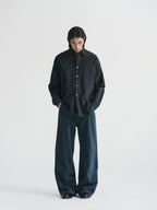 A model stands against a plain light gray studio background wearing a dark oversized button-front shirt with a subtle grid pattern and very wide-leg dark trousers from Inknots.