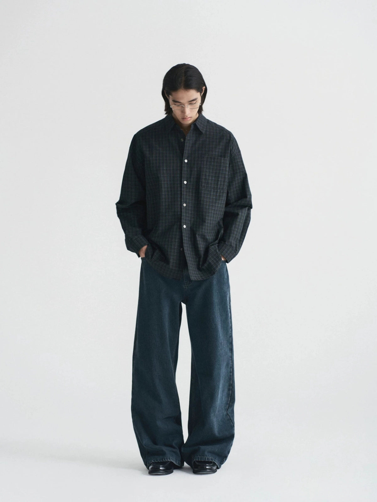 A model stands against a plain light gray studio background wearing a dark oversized button-front shirt with a subtle grid pattern and very wide-leg dark trousers from Inknots.