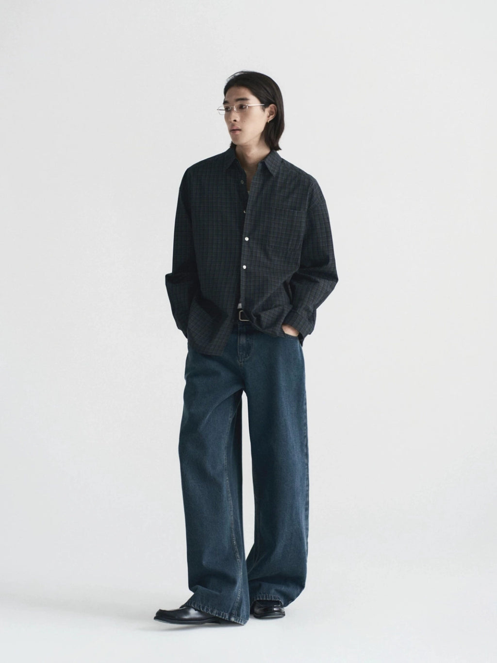 A model stands against a plain light gray studio background wearing a dark charcoal button-up shirt with a subtle grid pattern, paired with dark blue wide-leg jeans from Inknots.