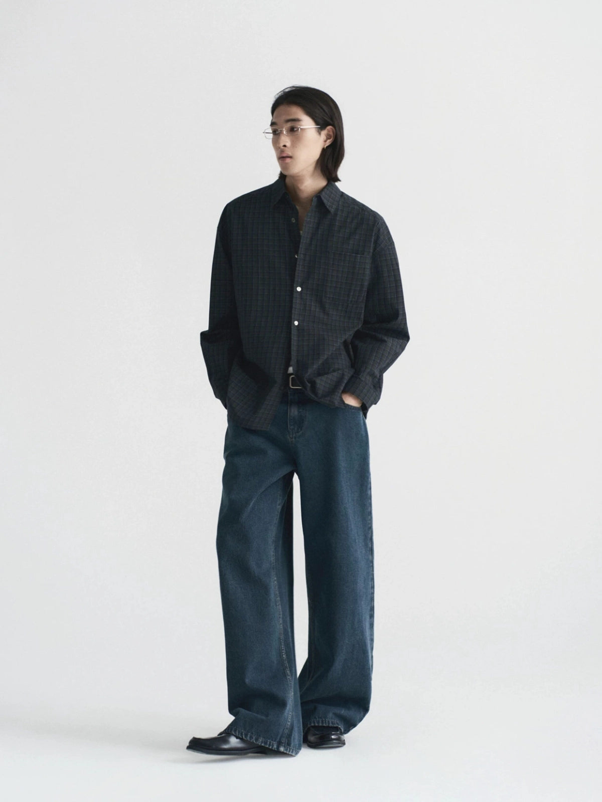 A model stands against a plain light gray studio background wearing a dark charcoal button-up shirt with a subtle grid pattern, paired with dark blue wide-leg jeans from Inknots.