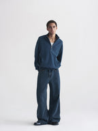 A man modeling a dark blue quarter-zip pullover and matching wide-leg jeans against a plain light gray studio background.