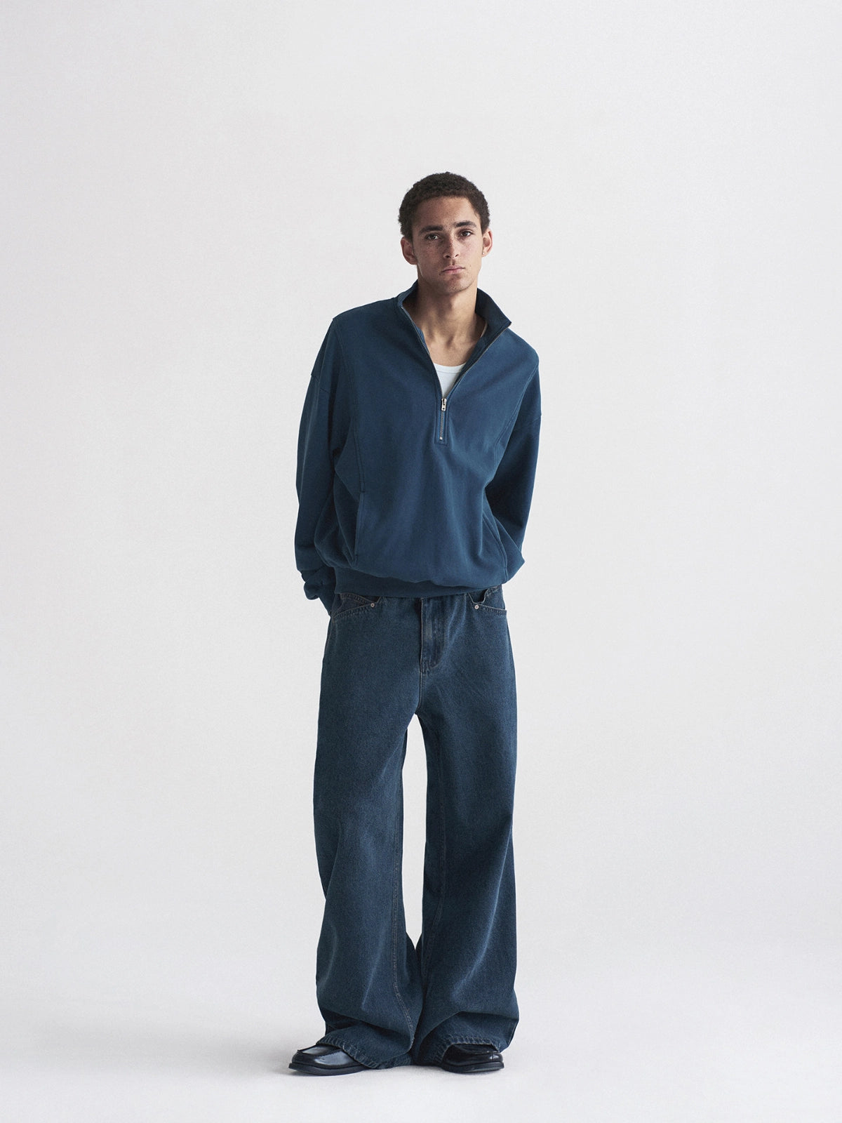 A man modeling a dark blue quarter-zip pullover and matching wide-leg jeans against a plain light gray studio background.