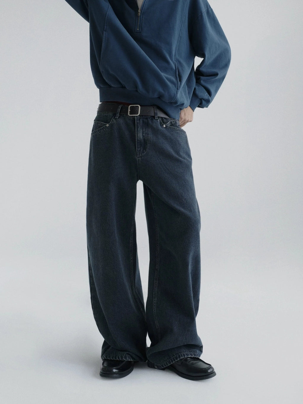 A person wearing dark wide-leg denim jeans with a brown belt, paired with a navy quarter-zip pullover from Inknots, photographed against a light gray studio background.